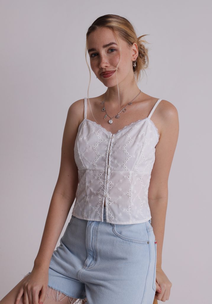 Cloud Dancer: looks com a cor de 2026 Menina branca, loira, veste blusa branca, com tecido laise, alças finas reguláveis, fechamento frontal por botões e comprimento cropped. Produto com a cor cloud Dancer, a cor de 2026